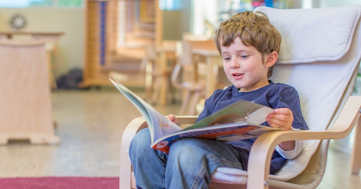 Young boy reading a book in a white chair.