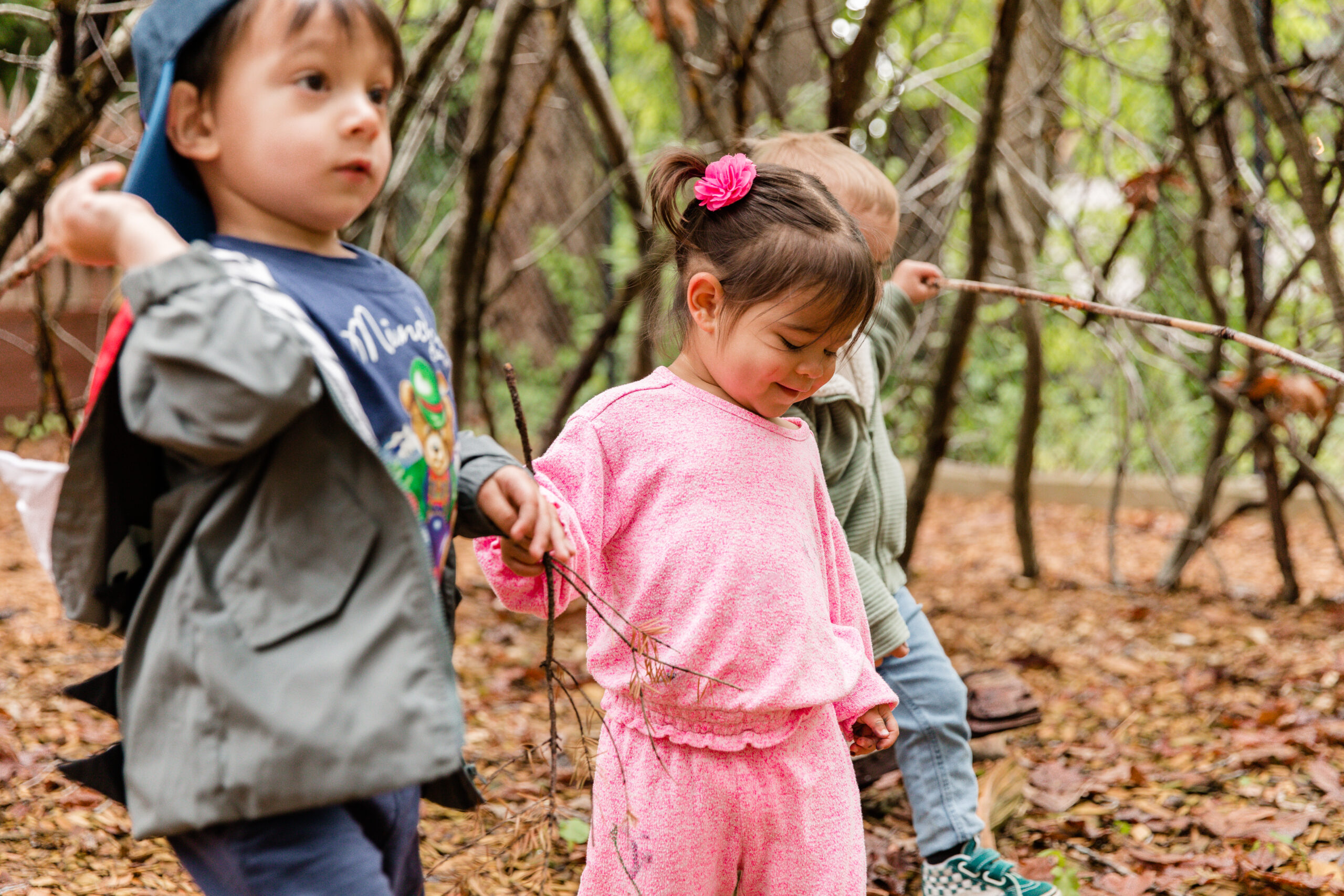 A young boy in a blue baseball cap an dgrey jacket walks through a wooded area with a young girl in a pink sweat suit.