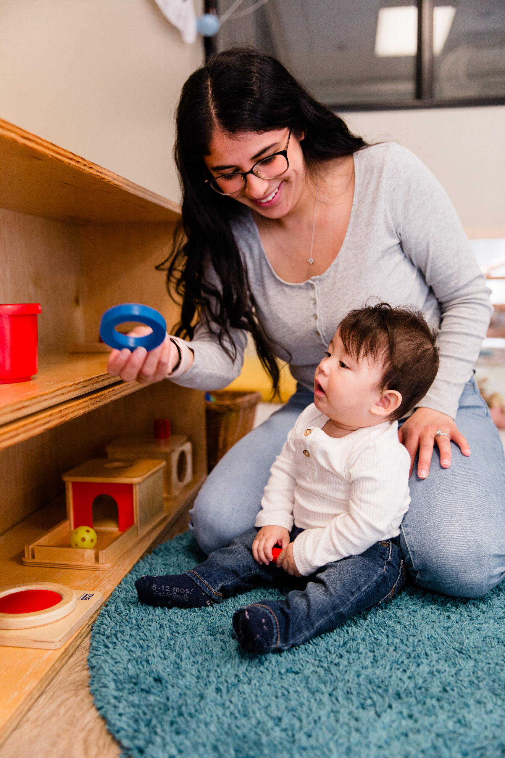 A mother with long black hair and glasses, wearing a grey top and blue jeans, props her baby with dark hair, a white top, and blue jeans on her legs.