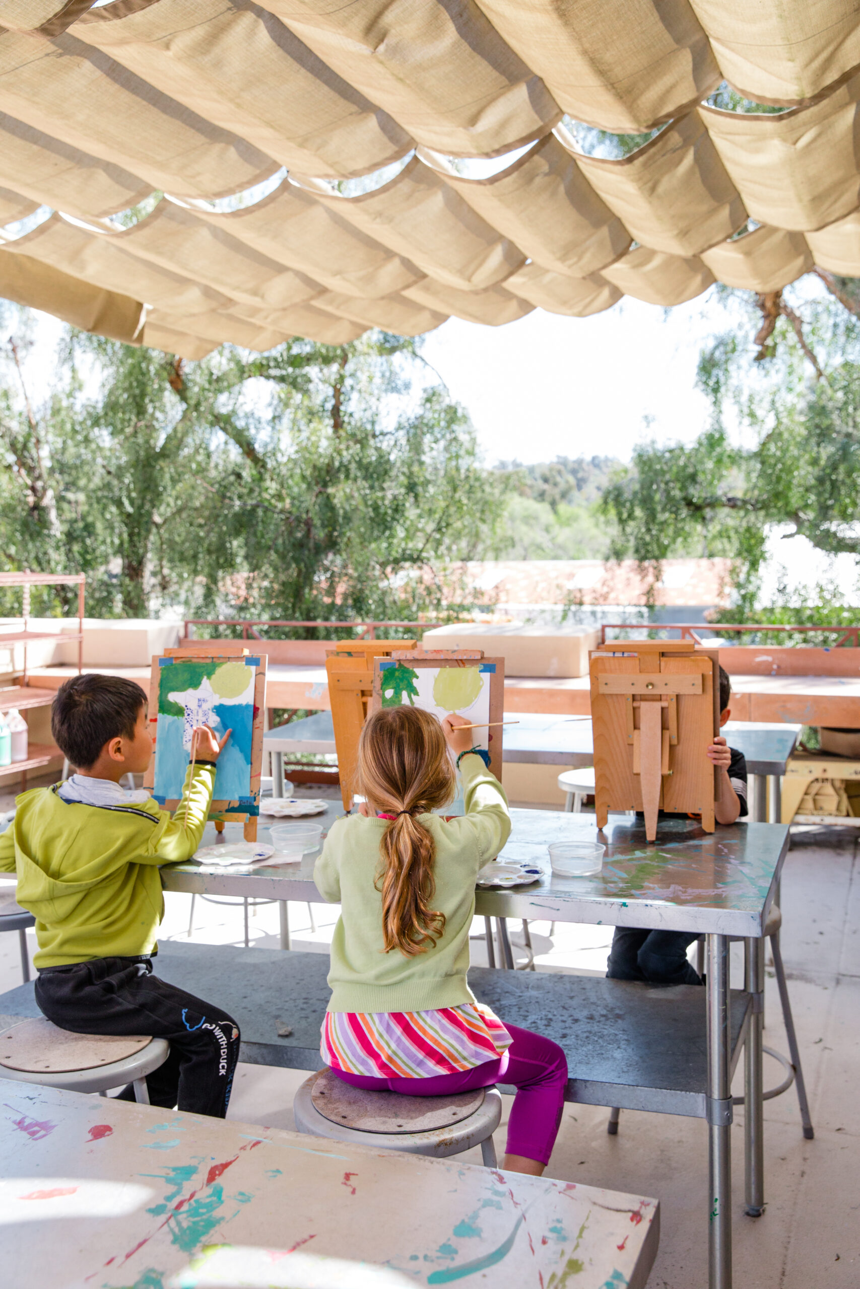 A girl with long hair, a light green sweater and a pink and white skirt sits at a metal table with an easel and paintbrush.