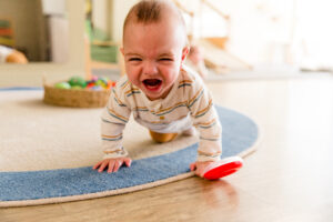 A baby crawls on all fours on a cream and blue rug. He is crying and wearing a white and grey striped onesie .