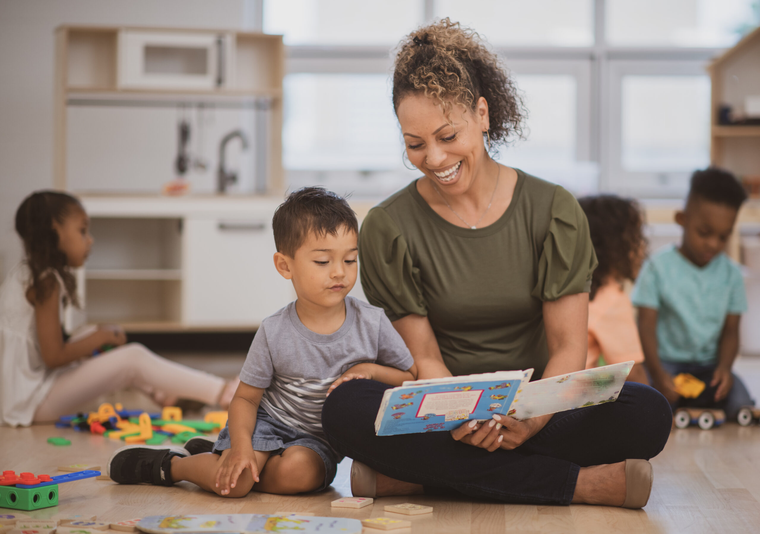 A preschool boy of Hispanic ethnicity sits next to his teacher as she is reading him a book on the classroom floor.story to a young boy with a grey shirt and black hair.